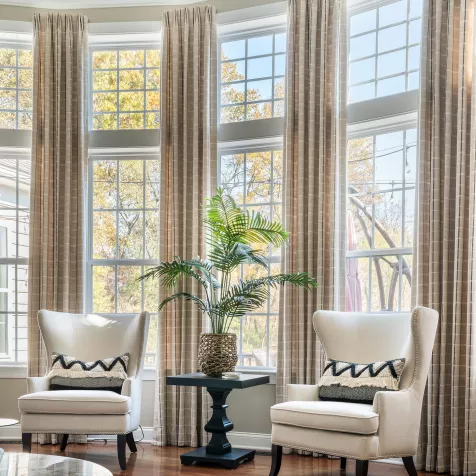 A bright room with two-story windows accented by long, tan, plaid drapery fabrics and two cream-colored wingback chairs, with a decorative plant between them.